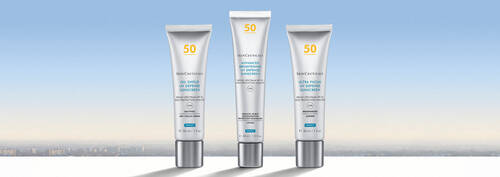 Three skincare tubes arranged on a white surface against a soft gradient background of blue and pale orange, depicting a skyline.
