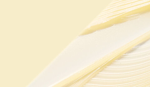 A close-up of a person's eye and hand, with the hand gently touching the skin near the eye. A small drop of liquid is visible on the skin. The background is plain and neutral.