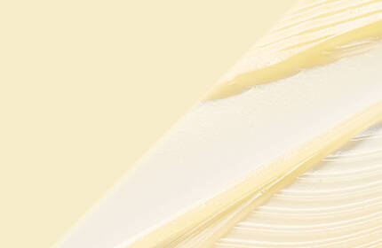 A close-up of a person's eye and hand, with the hand gently touching the skin near the eye. A small drop of liquid is visible on the skin. The background is plain and neutral.