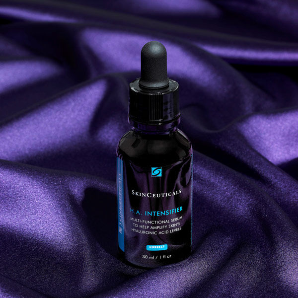 A bottle of SkinCeuticals H.A. Intensifier serum sitting on a purple satin background, featuring a dropper top and a sleek design.