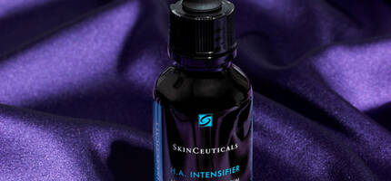 A bottle of SkinCeuticals H.A. Intensifier serum sitting on a purple satin background, featuring a dropper top and a sleek design.