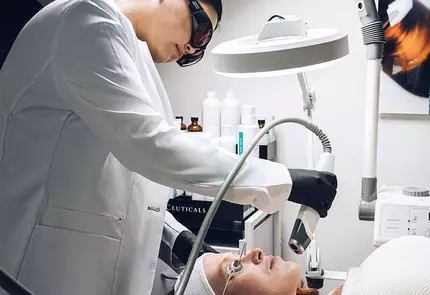Een huidtherapeut voert een behandeling uit op een patient in een schoonheidssalon, terwijl de patient op een behandeltafel ligt.