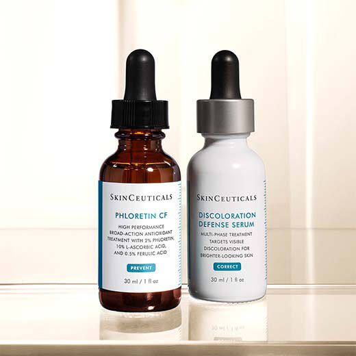 A photo featuring two skincare serum bottles from SkinCeuticals. One is a brown glass bottle labeled "Phloretin CF" and the other is a white bottle labeled "Discoloration Defense Serum." Each bottle contains 30 ml of product.