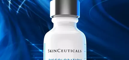 A white serum bottle with a dropper cap, labeled "SkinCeuticals Discoloration Defense Serum," against a vibrant blue abstract background. The text highlights that it targets visible discoloration for brighter-looking skin.