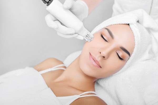 Een vrouw ondergaat een huidbehandeling met een microneedling-apparaat, terwijl ze ontspannen ligt met een handdoek om haar hoofd.