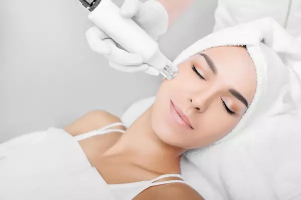 Een vrouw ondergaat een huidbehandeling met een microneedling-apparaat, terwijl ze ontspannen ligt met een handdoek om haar hoofd.