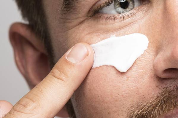 Een man met een baard die een gezichtscreme aanbrengt op zijn huid, terwijl hij met een vinger naar het product wijst.