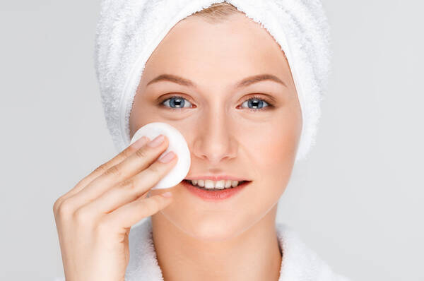 Een vrouw in een witte badjas en met een handdoek om haar hoofd, die een watje op haar gezicht aanbrengt.