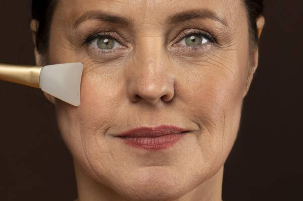 Een vrouw met een neutrale gezichtsuitdrukking die een schoonheidsproduct op haar wang aanbrengt met een spatel.