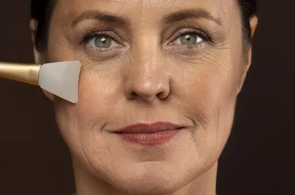 Een vrouw met een neutrale gezichtsuitdrukking die een schoonheidsproduct op haar wang aanbrengt met een spatel.