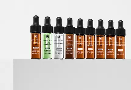 Een reeks van negen glazen pipetflessen met huidverzorgingsserums in verschillende kleuren, uitgestald op een witte ondergrond.