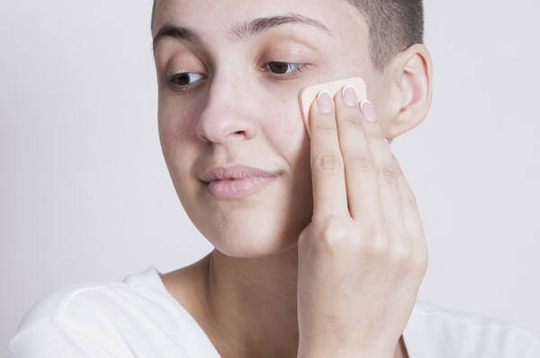 Een persoon met kort haar aan het aanbrengen van foundation op hun gezicht met een make-up sponsje.