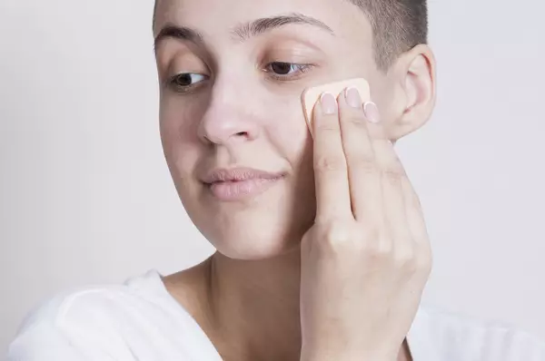 Een persoon met kort haar aan het aanbrengen van foundation op hun gezicht met een make-up sponsje.