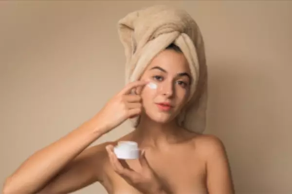 Een vrouw met een handdoek om haar hoofd brengt gezichtscreme aan op haar gezicht. Ze houdt een potje met creme in haar andere hand en kijkt recht in de camera. De achtergrond is neutraal van kleur.