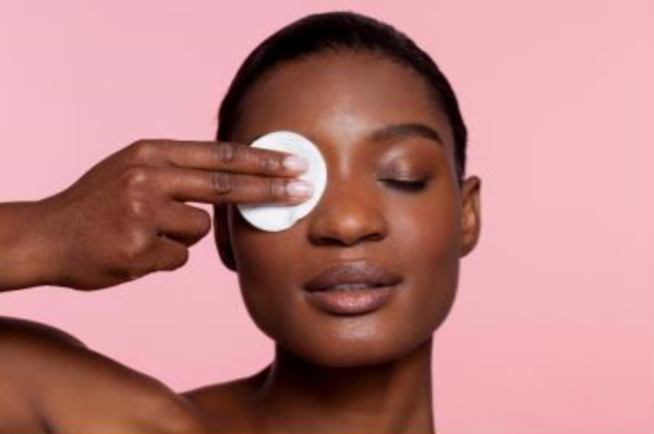 Een vrouw die een watje met gezichtsreiniger op haar gezicht aanbrengt, met een zachte, roze achtergrond.