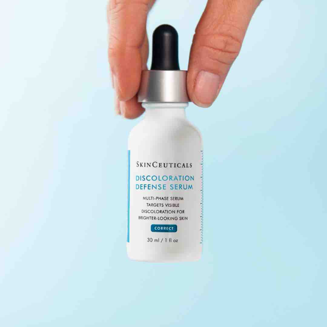 Een hand houdt een flesje van 30 ml SkinCeuticals Discoloration Defense Serum vast, met een druppelaar bovenop, tegen een lichte blauwe achtergrond.