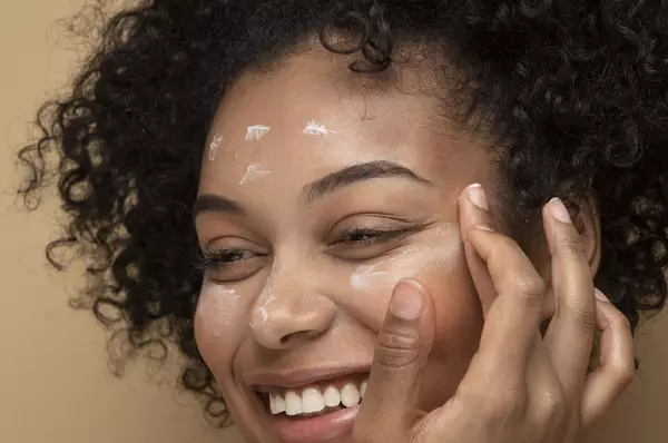 Een vrouw met krullend haar, die glimlacht terwijl ze een creme op haar gezicht aanbrengt, tegen een beige achtergrond.