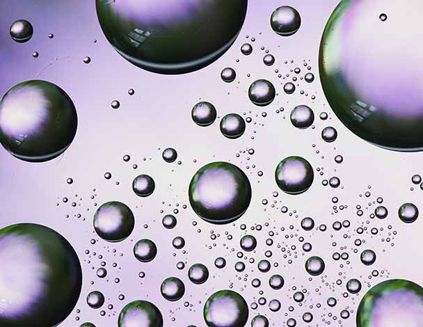 A close-up of iridescent bubbles of varying sizes against a soft purple background, creating a dynamic, airy effect.