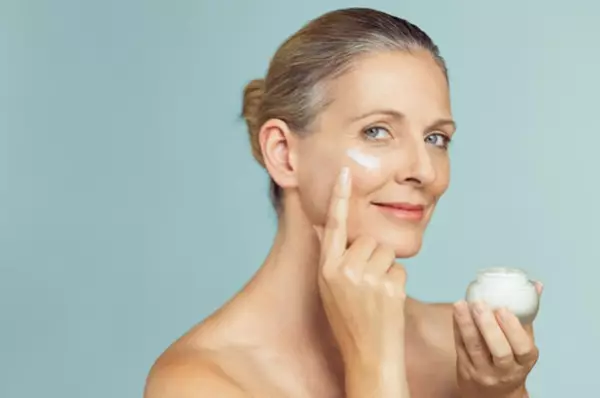 Vrouw zonder shirt met een glimlach, die creme aanbrengt op haar wang terwijl ze een potje creme vasthoudt.