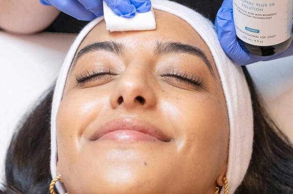Een vrouw met een witte hoofdband ligt ontspannen op een behandeltafel terwijl een persoon met blauwe handschoenen een gezichtsbehandeling aanbrengt met een watje.