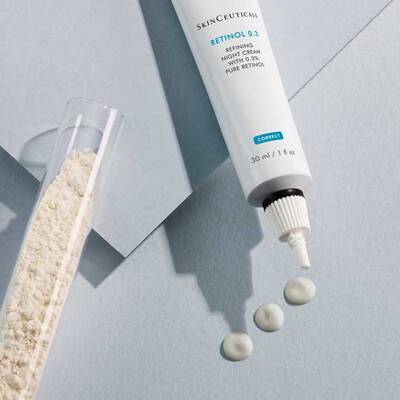 A tube of SkinCeuticals Retinol 0.3 cream is shown next to a small glass container with white powder, with three dabs of the cream on a light-colored surface.